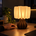 A modern table lamp with a textured, pleated white shade and a ribbed dark brown base, glowing warmly on a wooden desk next to a person's hand.