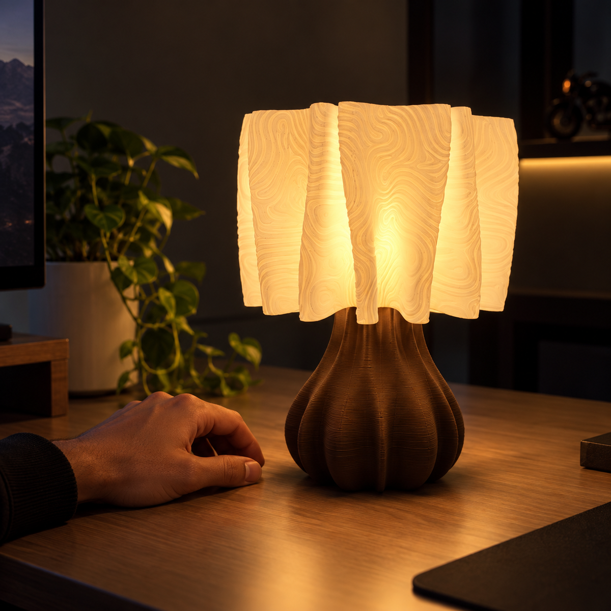 A modern table lamp with a textured, pleated white shade and a ribbed dark brown base, glowing warmly on a wooden desk next to a person's hand.